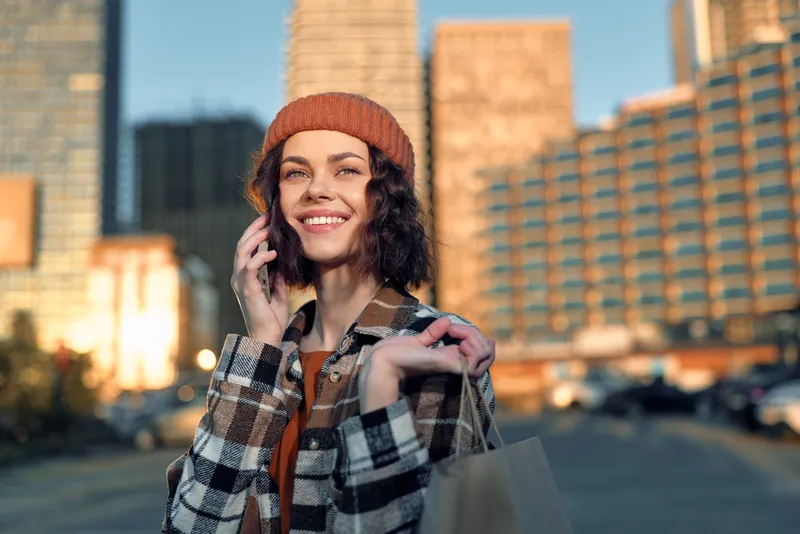 Woman with phone and shopping bag smile in city street at golden hour sunlight, candid lifestyle portrait exuding authenticity, mindful living and emotional storytelling.