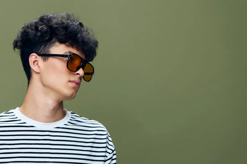 Young man with curly hair wearing sunglasses and a striped shirt posing against a solid olive green background in a casual style portrait.