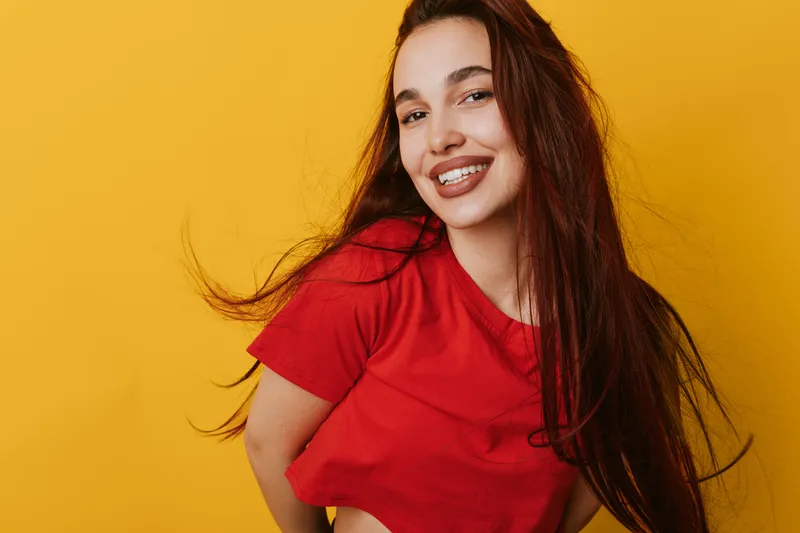 Young woman with long hair and red shirt smiling happily against yellow background. Portrait of cheerful female with natural makeup and confident expression in studio.