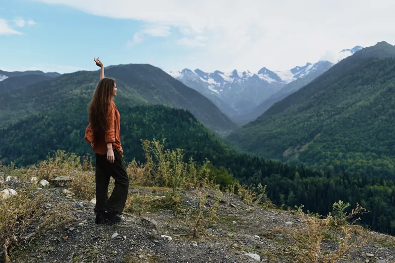 A hiker stands on rocky ground amid lush hills and towering snowy peaks, raising one arm toward the expansive valley for a dramatic outdoor landscape moment