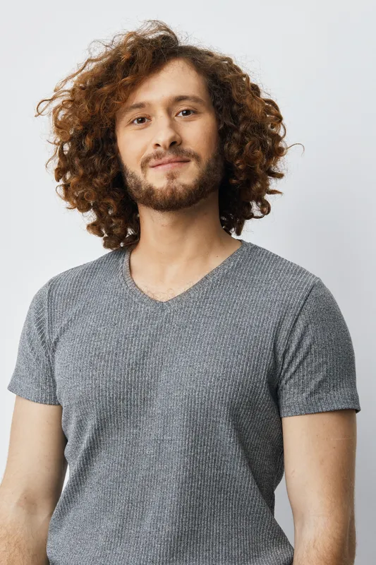 Happy young man with curly hair wearing a gray t shirt, showcasing a warm smile against a clean background, representing confidence and positivity