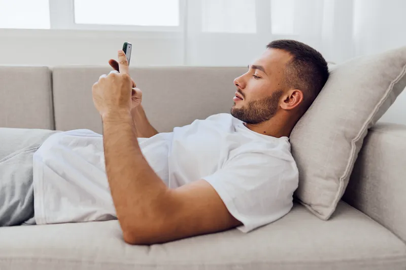 Relaxed man using smartphone on a cozy couch, showcasing modern lifestyle and technology in a minimalist setting with natural light.