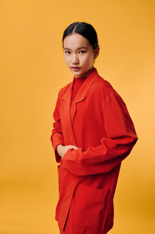 Young woman wearing red jacket standing with hands in pockets looking at camera on orange background. Portrait of confident female model with neat hairstyle and casual fashion style.