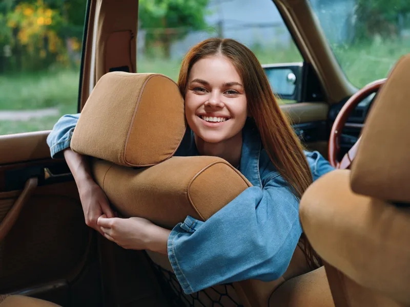 Young woman smiling inside a vintage car, surrounded by plush cushions, showcasing joy and comfort in a sunny outdoor setting, perfect for lifestyle themes.