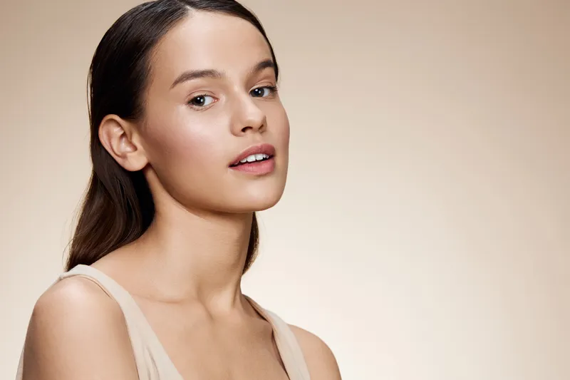 Natural beauty portrait of a young woman with long hair, neutral makeup, and soft expression against a light beige background for skincare and beauty promotions