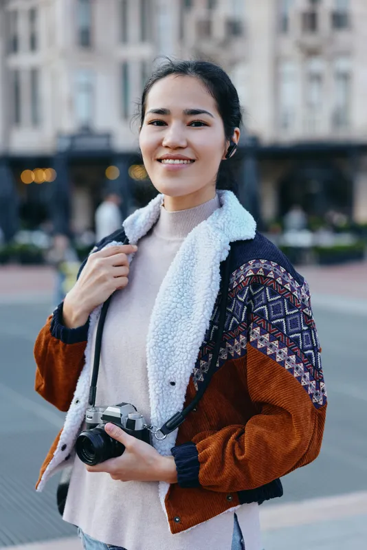 Camera in hand, woman on a busy street smiles for a candid photography scene in an urban outdoor setting, wearing a cozy jacket and scarf, embodying street style and lifestyle storytelling