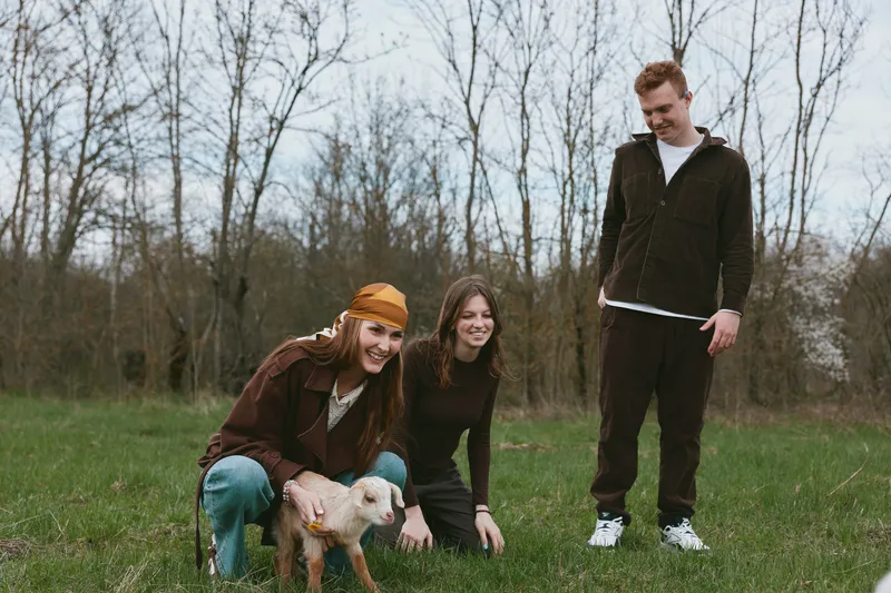 Happy trio outdoors with a playful goat kid, friends smiling by a grassy field, casual clothing, candid moment of companionship, warm sunlight, rustic vibe, peaceful rural scene, natural laughter and