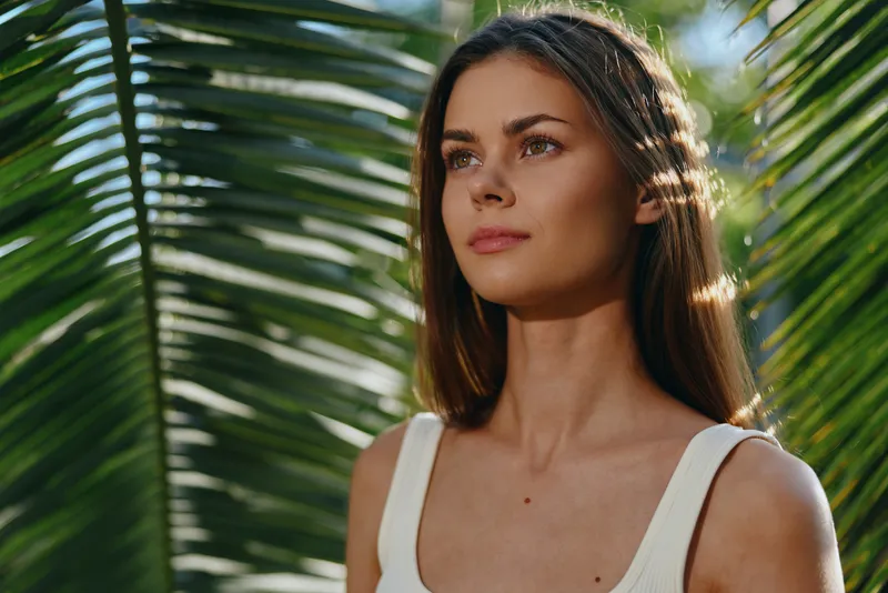 Young woman with long brown hair stands calmly against tropical palm leaves, wearing a white tank top in bright natural sunlight, expressing peaceful outdoor vibe.
