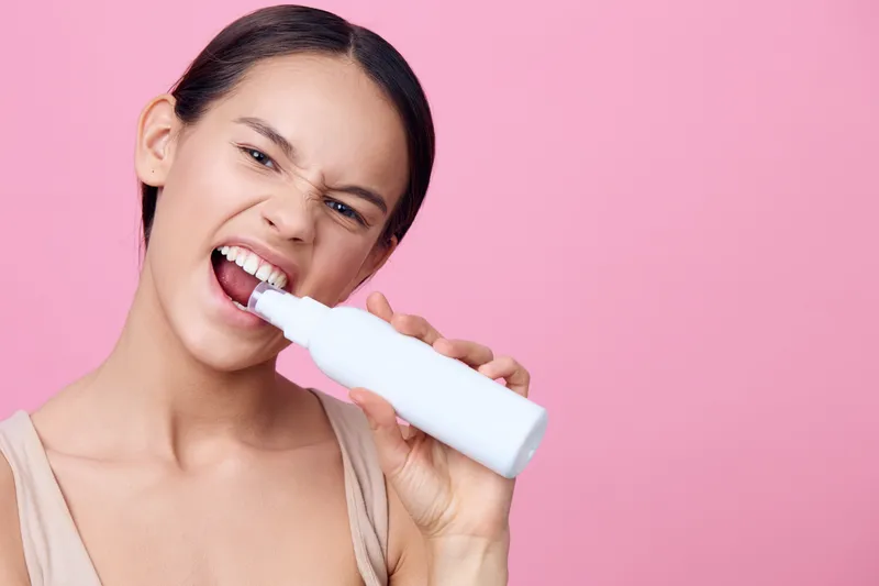 Young woman using a white dental care product, showcasing dental hygiene with a playful expression against a pink background