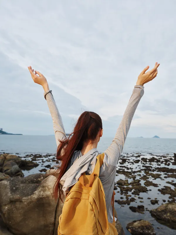 Young woman with long hair expressing joy against a scenic coastal background, celebrating freedom and nature, wearing casual attire with a backpack
