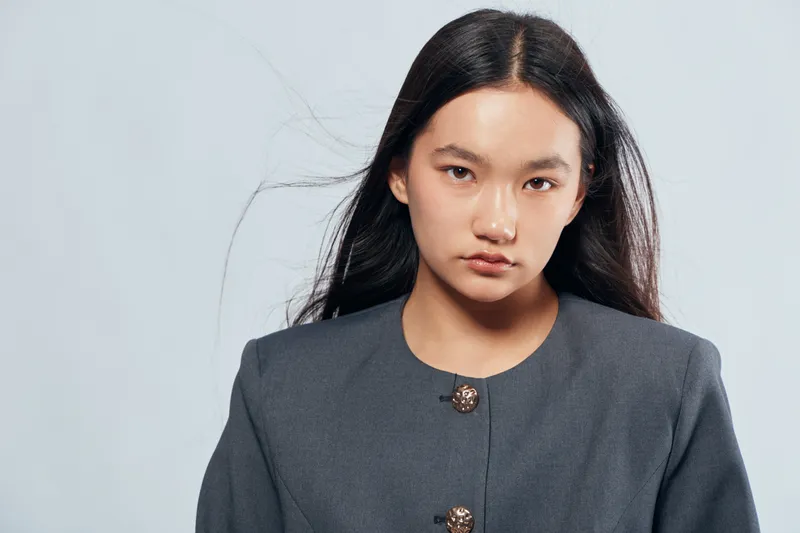 Young woman with long black hair and serious expression wearing gray blazer on light background, portrait of confident female with natural makeup and minimal accessories in studio.