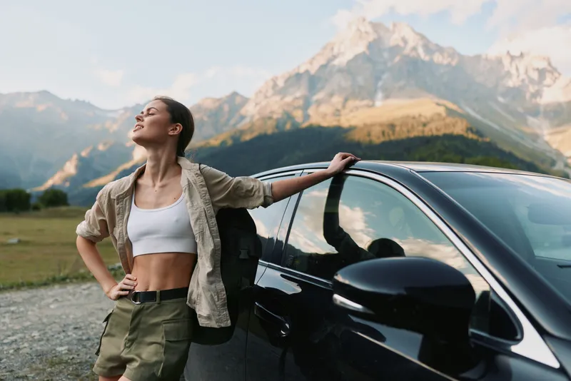 Smiling woman with backpack leaning on a car in a mountainous landscape, casual travel vibe, nature, adventure, and a relaxed, confident mood