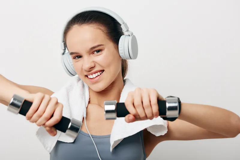Woman fitness, sport: Young woman fitness portrait holding dumbbells and wearing headphones and a towel, smiling confidently in a studio with white background, emphasizing strength and active