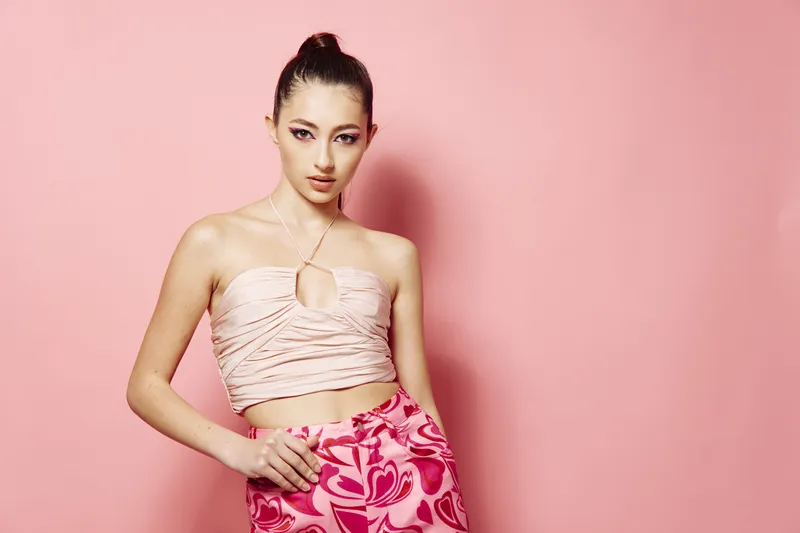 Fashionable young woman with sleek hair poses against a pink wall wearing a delicate top and patterned skirt, showcasing edgy beauty and creative fashion style.