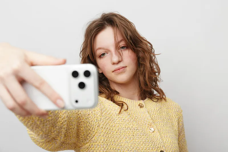 Young woman with curly hair wearing yellow sweater takes selfie with smartphone on white background. Casual style, natural expression, social media concept, modern technology use.