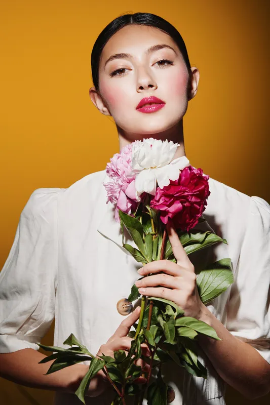 Young woman with pink and white flowers looks confidently into the camera against warm orange backdrop. Soft natural makeup enhances spring fashion and creative beauty concept.