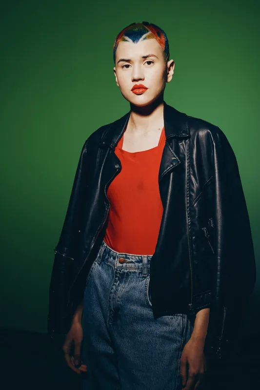 Young woman with short colorful hair wearing a black leather jacket and red top standing against green background in casual fashion style, confident and modern look.
