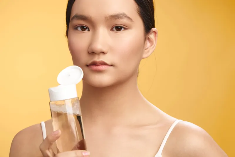asian woman holds a clear bottle of skincare product, looking softly at the camera with a calm, confident expression in a bright studio setting.