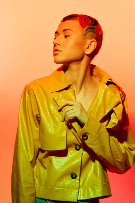 Woman with unique hairstyle wearing a bright yellow jacket, looking thoughtfully to the side against a warm orange background with dramatic lighting and shadow play.