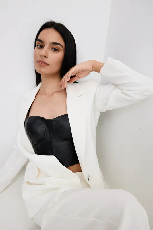 young woman in white suit and black corset posing indoors against white wall, showcasing modern fashion and confident style in minimalist setting