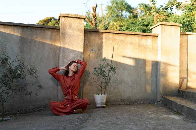 A woman in a red dress sits on a sunlit courtyard ground, stretching with her arms raised and a relaxed, contemplative expression, warm shadows creating a calm mood