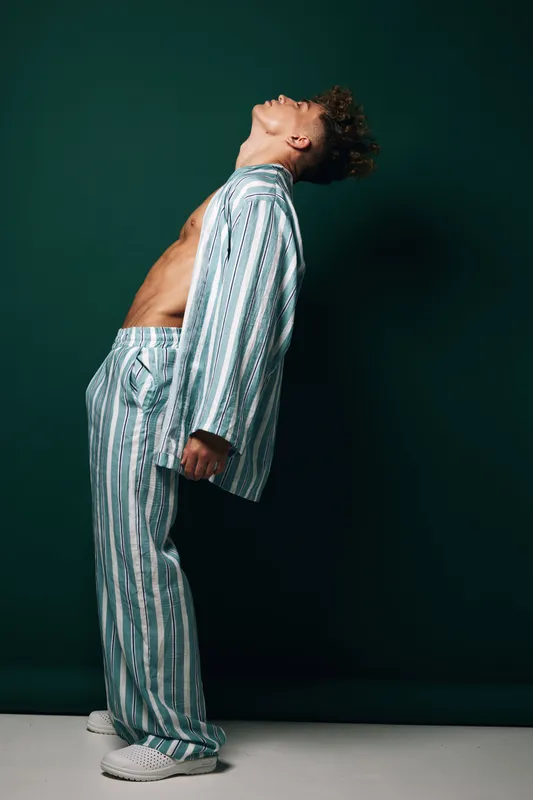Young man in striped pajamas leaning backward with bare chest against dark green background, casual pose and relaxed mood in studio lighting for fashion or lifestyle concept.