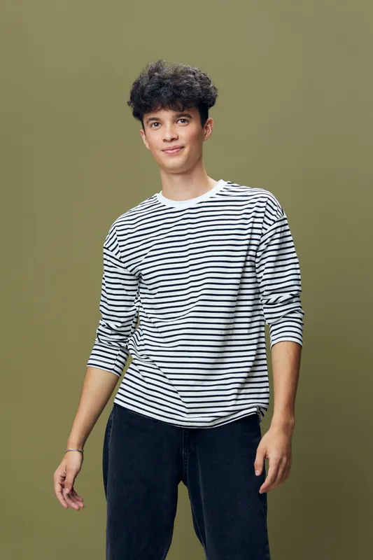 young man in striped shirt standing with relaxed posture against plain olive background, casual clothing, curly hair, youthful expression