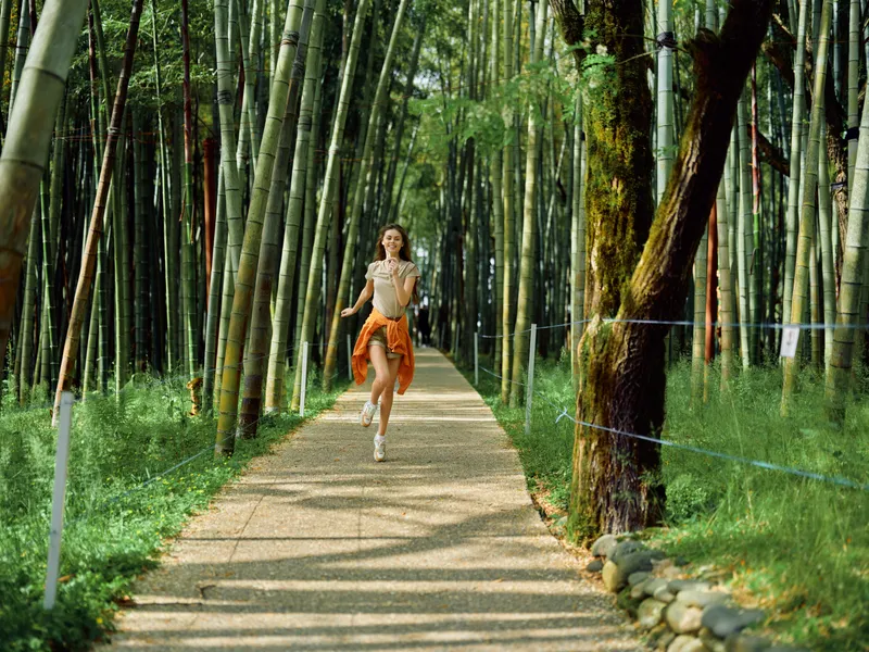 woman running along a sunlit bamboo path in a serene forest, jogging for fitness and joy. Young runner in athletic outfit on gravel trail surrounded by green nature and trees