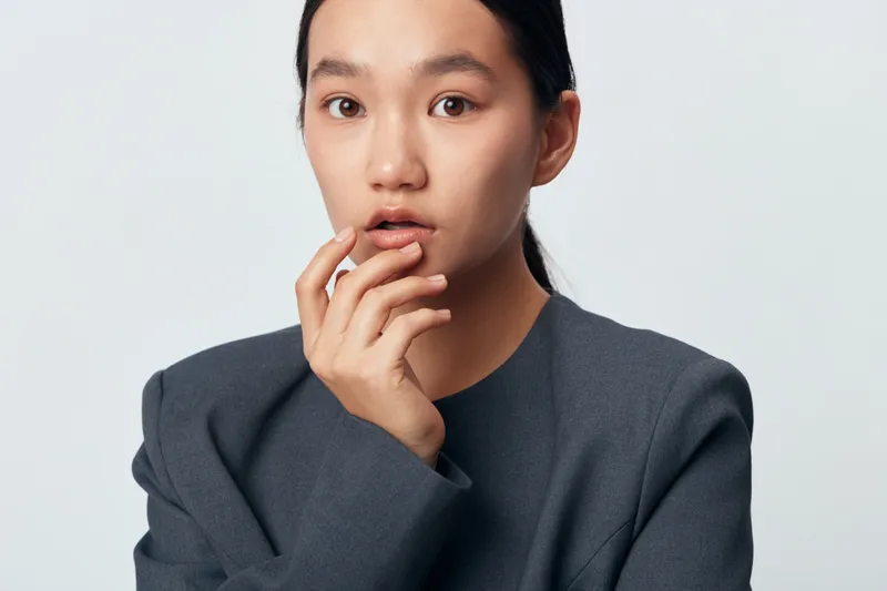 Portrait of young woman touching lips with fingers in gray blazer against neutral background, expressing thoughtful and calm mood in studio lighting with natural makeup and tidy hairstyle.