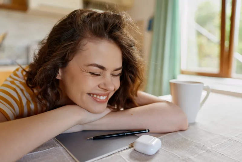 Smiling woman rests her chin on her arms at a desk, with a pen and tablet nearby. Morning light through a window creates a cozy, welcoming, natural home workspace mood, quiet and calm.