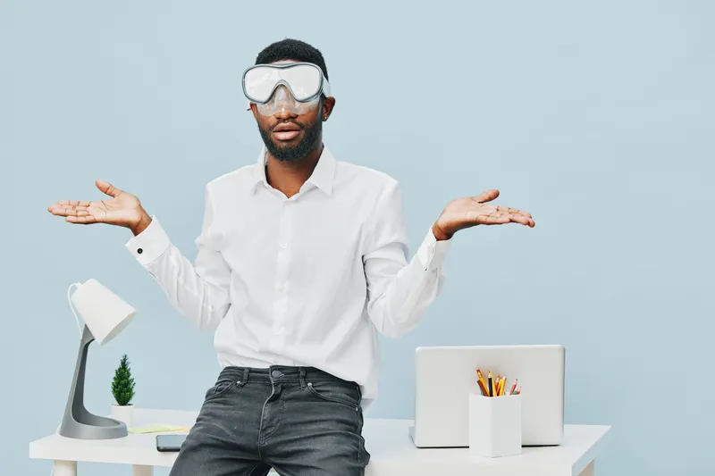 Confused man with glasses shrugging shoulders indoors, workspace with computer and lamp, casual look, light blue background, expression of uncertainty, modern office environment, lifestyle concept, re