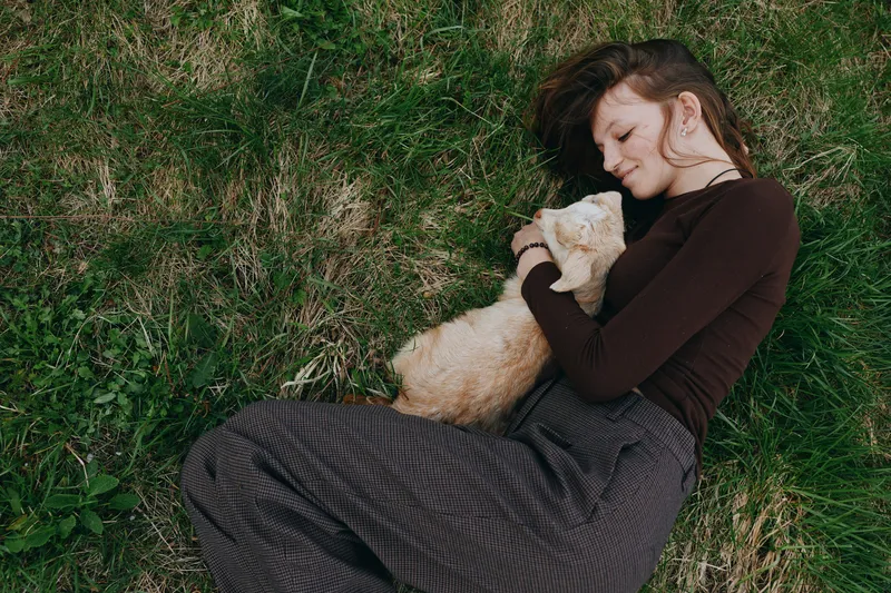Relaxed woman lying on grass, cuddling a fluffy animal, enjoying outdoor relaxation, calm mood, serene lifestyle, nature light, field and countryside vibes, cozy moment with pet.