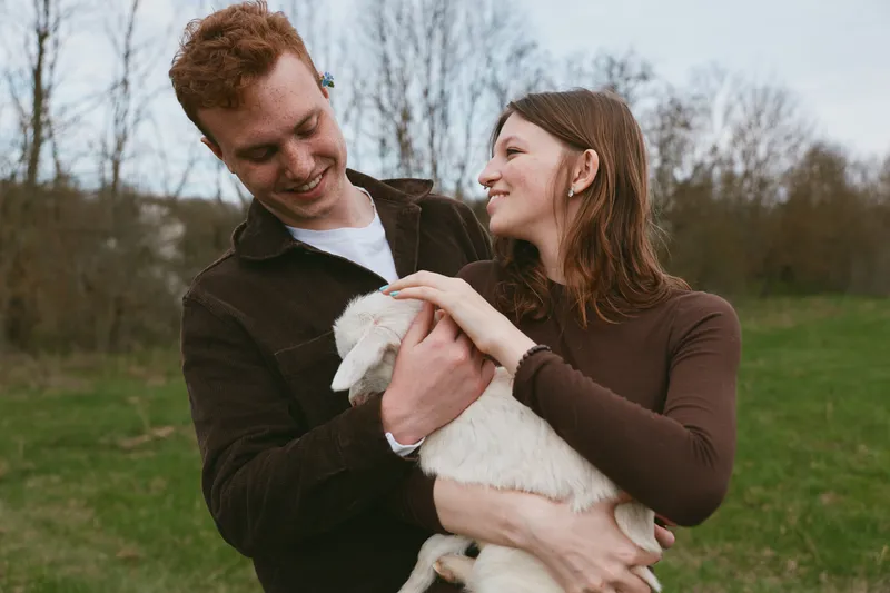 couple, goat, kid, outdoors, cheerful scene of a smiling pair gently holding a playful goat kid in a sunlit field, natural colors, candid moment, warm connection, lifestyle emphasis