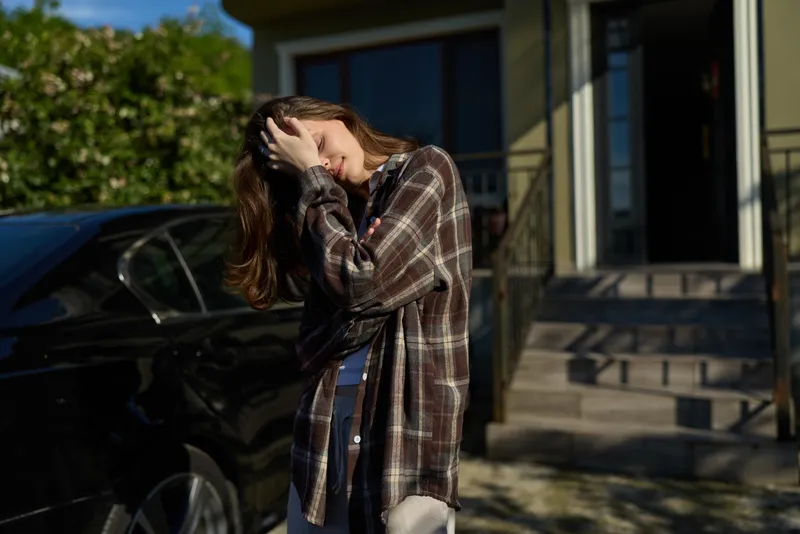 Young woman with long hair adjusts her hair beside a car outside a house on a sunny day
