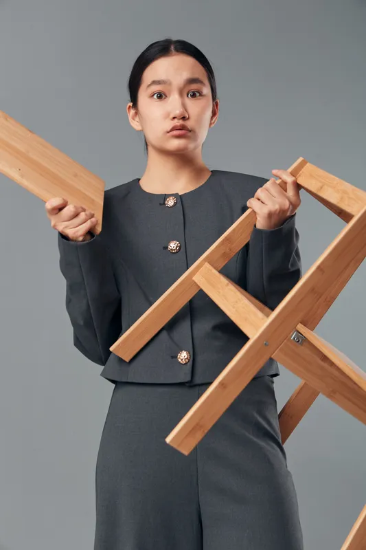 Woman with focused expression holding wooden chair frame in hands, dressed in dark formal outfit isolated on grey background for craftsmanship concept and furniture making.