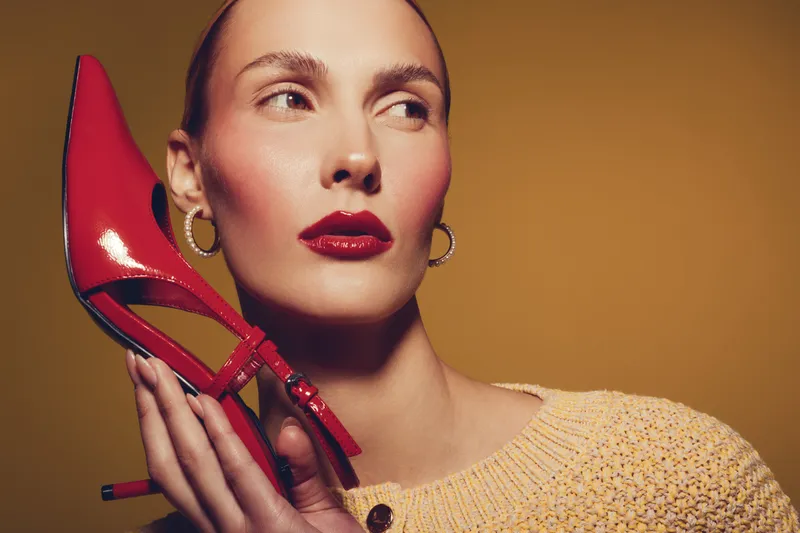 Stylish woman with glossy red lipstick holds red high heel shoe near face, wearing yellow knitted sweater and gold hoop earrings. Fashion concept with bold makeup and accessories.