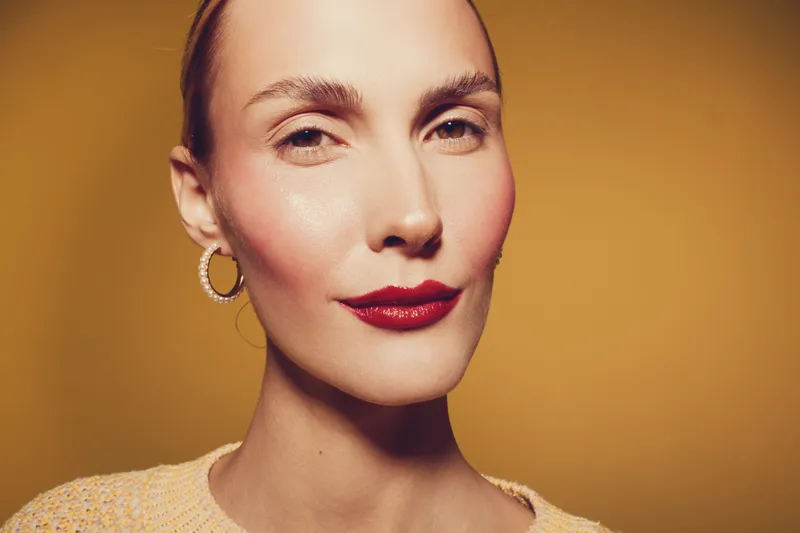 Close-up portrait of confident woman with red lipstick and hoop earrings on yellow background. Concept of beauty, fashion, and makeup with warm studio lighting.