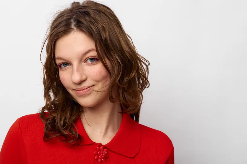 Young woman with wavy brown hair wearing a red sweater smiles gently against a plain white background. Concept of natural beauty, casual style, and positive mood portrait.