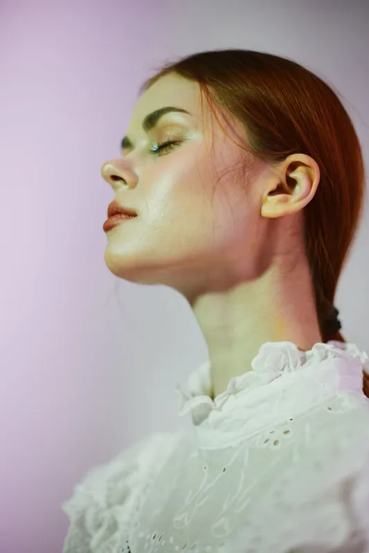 Ethereal portrait of a young woman with red hair in vintage lace blouse, expressing calmness and serenity against a softly lit pastel background