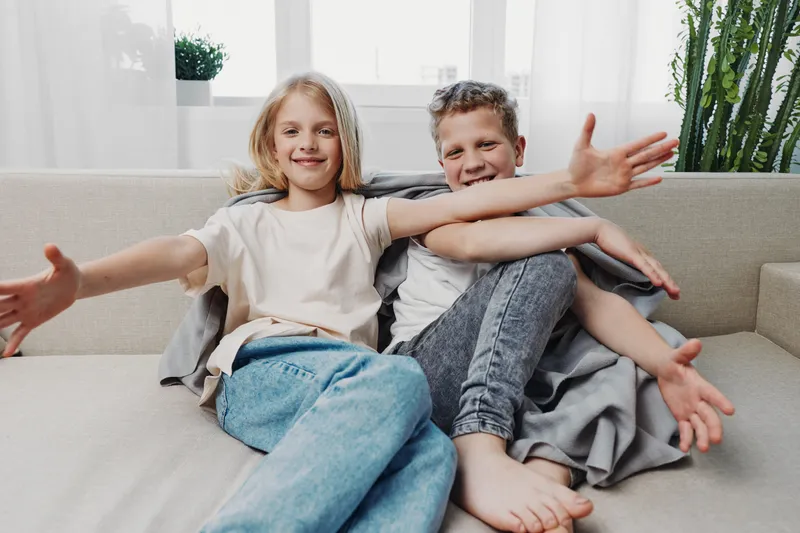 Two happy children sitting on a couch with open arms in a bright living room, smiling and enjoying casual home moment.