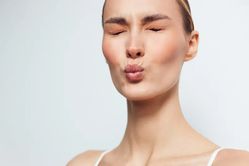 A young woman with clear skin and tied back hair puckers her lips and closes her eyes against a plain light background. Concept for skincare, beauty, and natural expression.