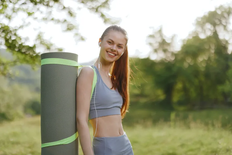 Smiling young woman in athletic wear holding a yoga mat outdoors, embodying fitness, wellness, and a healthy lifestyle in nature with a vibrant background