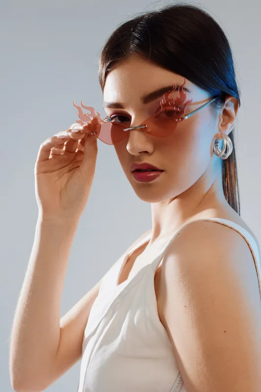 Young woman in white top wearing stylish pink sunglasses and hoop earrings looks confidently at camera. Fashion portrait with beauty concept in soft lighting.