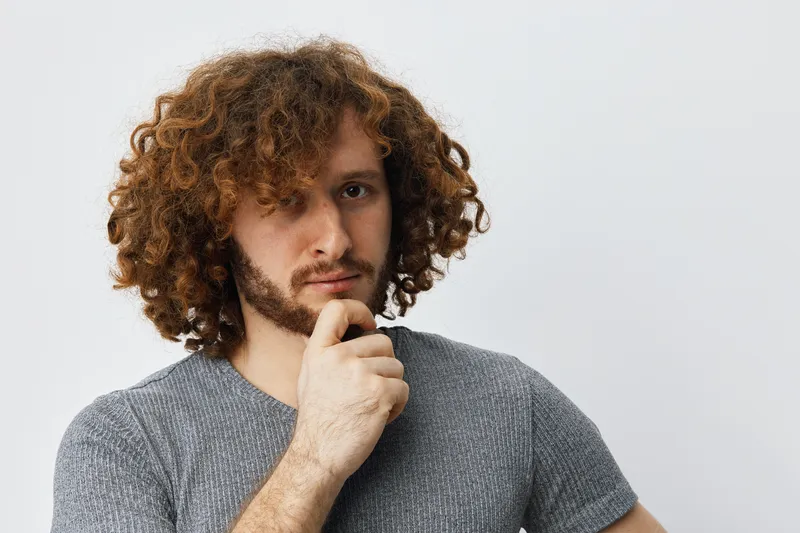 Thoughtful young man with curly hair in a gray shirt, looking pensive against a neutral background, representing introspection and self reflection
