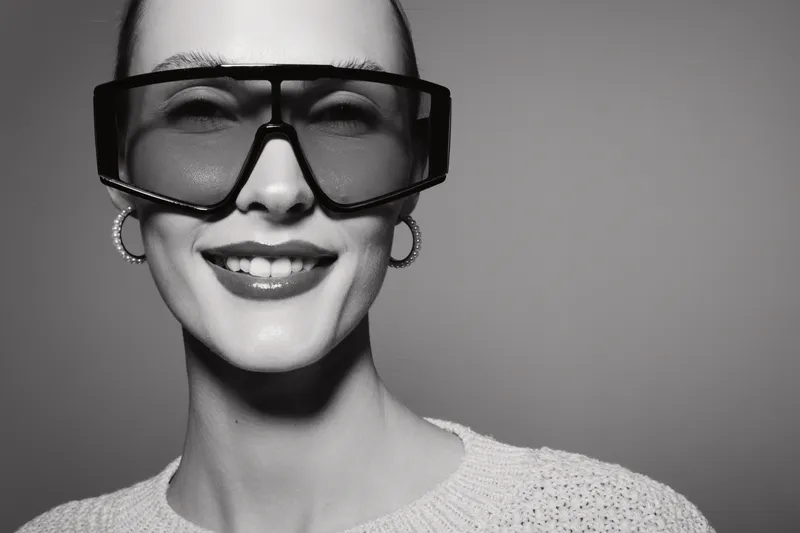 Smiling Caucasian woman with pale skin wearing stylish oversized sunglasses and earrings in monochrome studio portrait.