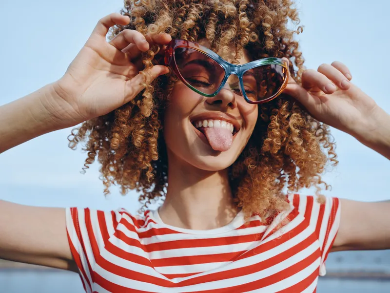 Playful portrait of a young person with curly hair wearing colorful sunglasses and a red and white striped shirt, sticking out tongue against a bright blue sky.