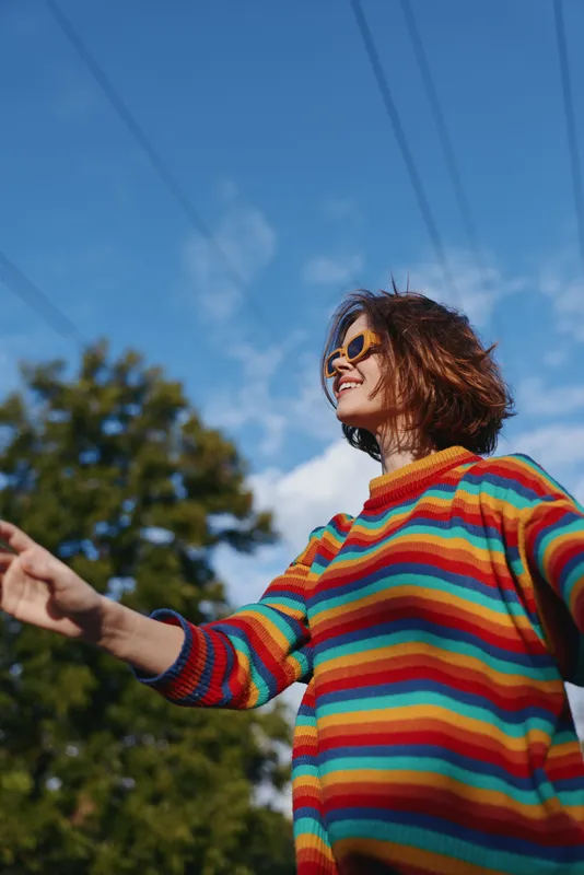 Woman in colorful sweater wearing sunglasses and smile outdoors under blue sky, portrait of a happy young adult enjoying a sunny day among trees, casual fashion and freedom.