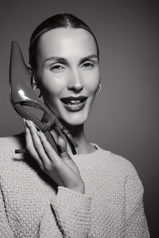 Young caucasian woman with fair skin holding shoe near ear smiling, dressed in knitted sweater, studio portrait in black and white with soft lighting.