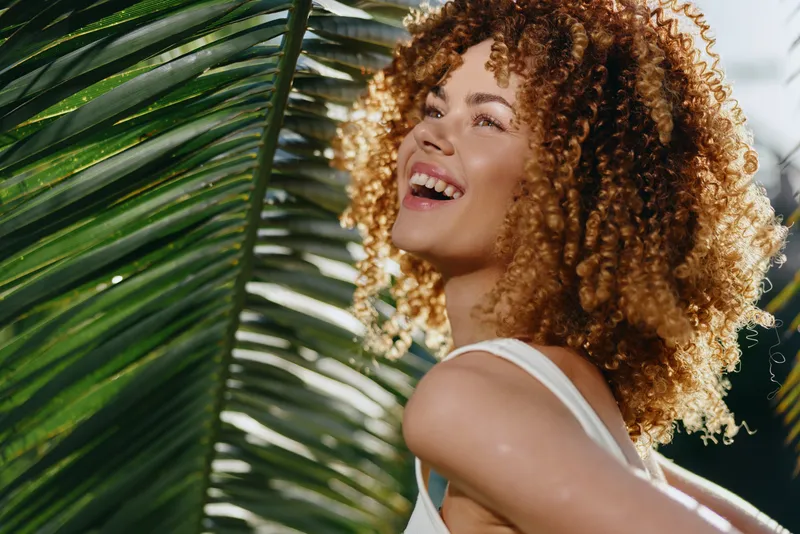 Smiling woman with curly hair enjoying sunlight outdoors near tropical palm leaves, expressing happiness, warmth, and natural beauty on a sunny day.