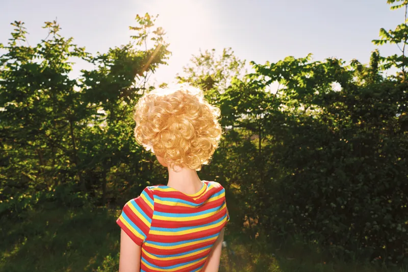 Young woman with curly blonde hair stands in a sunlit field wearing a striped shirt viewed from behind, conveying carefree summer mood and natural outdoor simplicity.